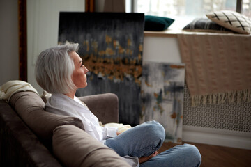 Side view of peaceful middle aged female with gray hair and in trendy clothes sitting on cozy sofa...