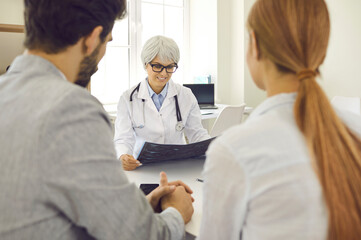 Obraz premium Mature female doctor in hospital advises expectant parents on planning pregnancy. Smiling health worker examines X-ray of married couple sitting with their backs to camera. Selective focus.