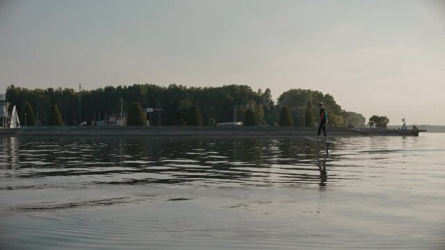 Man riding on a hydrofoil surfboard on large lake at golden sunset. Shot of man riding a hydrofoil surfboard near lake shore and marina. High quality 4k footage