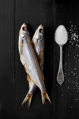 Two air-dried sabrefish on black wooden surface with spoonful of salt