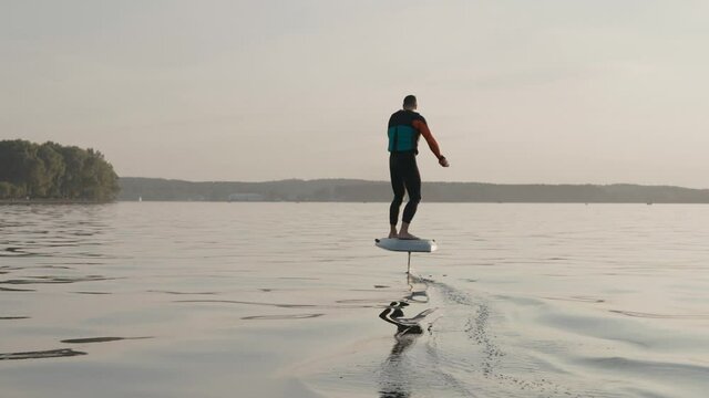 Man riding on a hydrofoil surfboard on large lake at golden sunset. Shot of man riding a hydrofoil surfboard near lake shore. High quality 4k footage