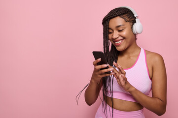 Beautiful African American sports woman in pink sportswear and wireless headphones browses web pages on internet in smartphone, smile with cheerful toothy smile, isolated against colored background