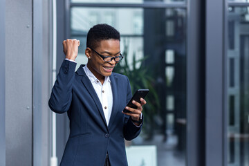 Happy and successful business woman rejoices in winning and happy day, celebrates, african american smiling holds phone in hands