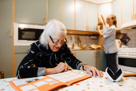 Senior woman solving sudoku while female nurse doing chores in kitchen