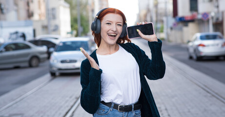 A beautiful, attractive and happy redhead Caucasian girl with headphones listening to music, using a smartphone and singing a song.