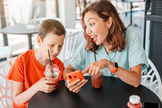 Mother Shows Her Son Funny Memes On Smartphone Screen, But Her Son Does Not Understand Her And Frowns. Generational Difference And Communication Problems