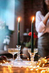 Lifestyle trendy Christmas table setting with blurred woman on background. Festive New Year decor for holiday romantic dinner for two with champagne, candles, glittering lights. Selective soft focus  