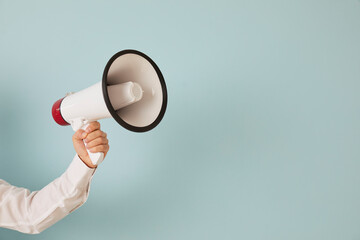 Attention please. Unknown man's hand on light blue background holds loudspeaker which is symbol of...