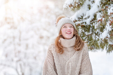 A young woman in a knitted hat and a warm sweater has a funny snowflake on her nose. The girl stands in the forest and laughs, looking up. The joy of the arrival of winter, the first snow concept