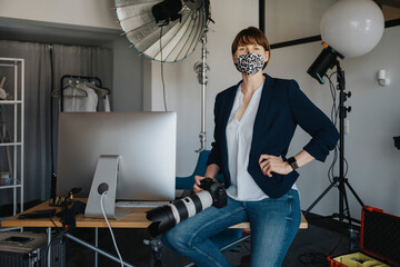 Female photographer sitting with camera on desk in studio during pandemic