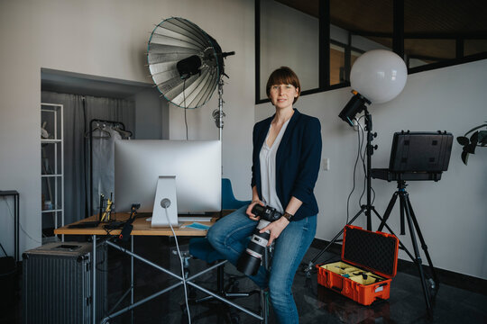 Confident female photographer holding digital camera while sitting on desk at studio