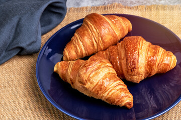 Three fresh croissants on a blue plate on a raffia tablecloth next to a napkin