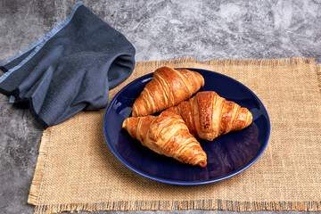 Three fresh croissants on a blue plate on a raffia tablecloth next to a napkin
