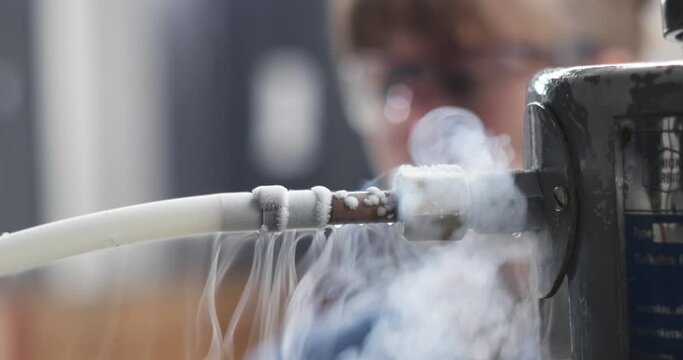 Unrecognizable female science lab technician supervising the transfer of liquid nitrogen from a storage tank through a pipe and valve. Real time, shallow depth of field