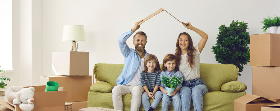 Portrait Of Happy Family With Kids Sitting At Home On Sofa Holding Cardboard Roof Over Their Heads. Young Family Feels Happy And Protected In Their New Apartment. Insurance Concept. Banner.