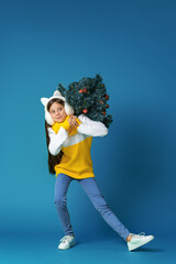 happy teenage girl in knitted sweater, full-length, wearing fur headphones on her head, with Christmas tree decorated with red Christmas balls on her shoulder, walks on blue background. Dynamic image.
