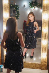 little girl in her mother's dress and high-heeled shoes is standing at home in living room. baby wants to be big and she likes way she looks. child in an adult dress and shoes is reflected in mirror