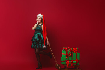 attractive young girl in a green dress and Santa Claus hat, riding a sleigh, delivering New Year's gifts, isolated red background. New Year, Christmas sale. Copy space. In full growth