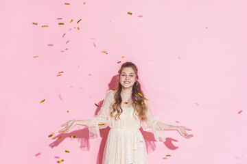Obraz na płótnie Canvas Merry Christmas. A beautiful attractive girl with wavy hair in a light shiny dress dancing under golden falling, flying confetti on a pink background in the studio, filmed with harsh light. Copy space