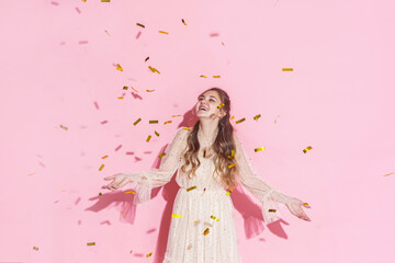 Obraz na płótnie Canvas Merry Christmas. A beautiful attractive girl with wavy hair in a light shiny dress dancing under golden falling, flying confetti on a pink background in the studio, filmed with harsh light. Copy space