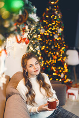 Attractive girl sitting on a sofa in a cozy room, with a mug in her hands, in a white knitted sweater. the beauty smiles, looks away, the home interior was decorated for Christmas. blinding light.