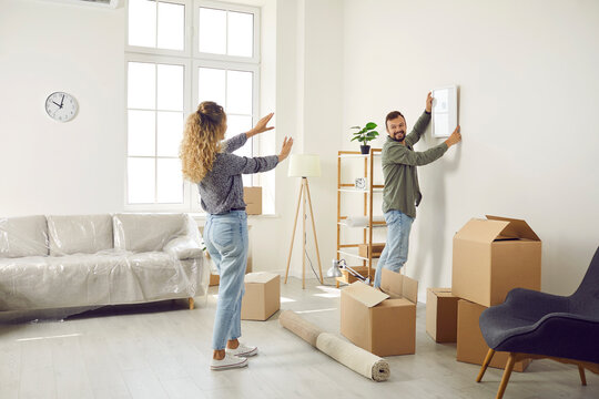 Happy married couple who recently bought house are unpacking stuff and decorating new home. Young man and woman are making living room cozy and choosing place on wall to hang painting or family photo