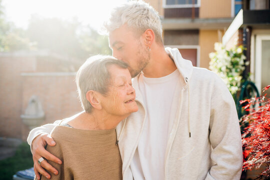 Young man kissing grandmother forehead while standing with arm around at backyard