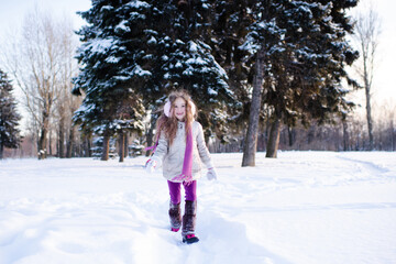 Cute funny kid girl 5-6 year old walk in snowy park over nature background outdoors. Little child wear winter jacket, scarf and headphones outside. Childhood.