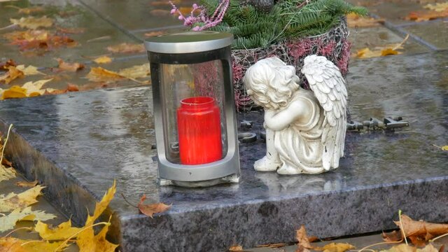 Red memorial candle burning with fire on rainy day on tombstone in cemetery autumn, rainy day. Grave with tombstone on raindrops falling, against abackground of yellow foliage next to sad angel statue