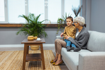 Little grandson talking with attentive happy grandmother, sitting on cozy sofa at home, smiling grandma with little boy having pleasant conversation, grandchild with granny having fun together