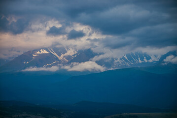 Atmospheric mountain landscape with great snowy mountains in low clouds and sunset cloudy sky. Awesome minimal alpine scenery with big glacier on high mountain ridge in low clouds in blue white colors