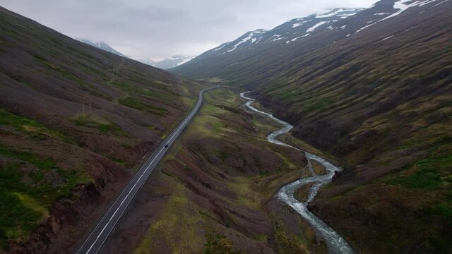 Aerial shot flying over a car driving through valley in Iceland. Dramatic overcast day with snowy mountains. 
