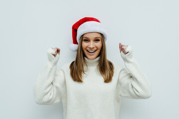 Young woman wearing a Santa hat over white background very happy and excited making winner gesture with raised arms, smiling and screaming for success.