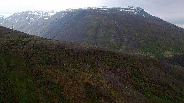 Aerial shot flies over a hill revealing Icelandic landscape beyond. Dramatic valley with river and snowy mountains.