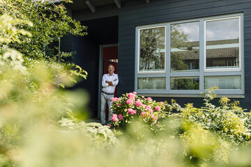 Man with arms crossed leaning on wall outside house during sunny day