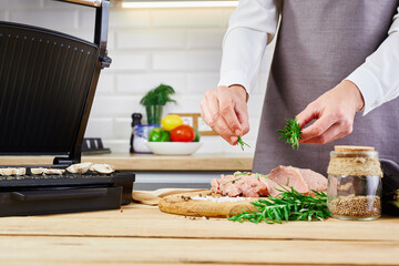 Woman cooking meat steak on kitchen. Female chef sprinkles fresh raw meat on a kitchen background with rosemary and salt. Electric grill and meat on wooden table.