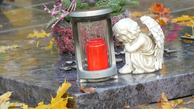 Red memorial candle burning with fire on rainy day on tombstone in cemetery autumn, rainy day. Grave with tombstone on raindrops falling, against abackground of yellow foliage next to sad angel statue