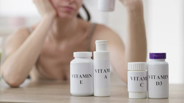 Blurred thoughtful woman sitting at table with row of vitamin bottle choosing pills. Unrecognizable Caucasian young slim lady thinking on dietary supplements indoors. Medicine and beauty concept