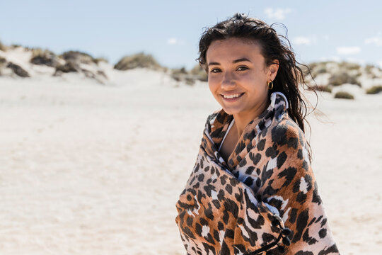 Beautiful smiling woman wrapped in scarf standing at beach on sunny day