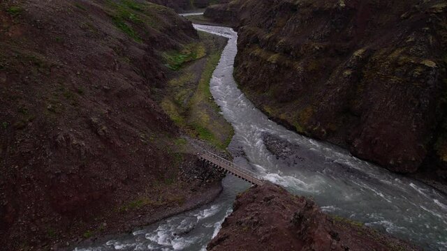 Icelandic aerial begins down and pans up to reveal mountain valleys. River flows below with a bridge, on a cloudy foggy day.