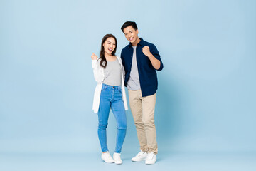 Full lenght portrait of young smiling Asian couple holding each other and clenching fists in isolated light blue studio background