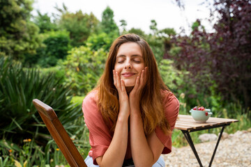 Smiling woman with eyes closed touching face while sitting on chair in garden
