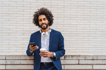 Male entrepreneur with mobile phone and disposable cup standing in front of wall