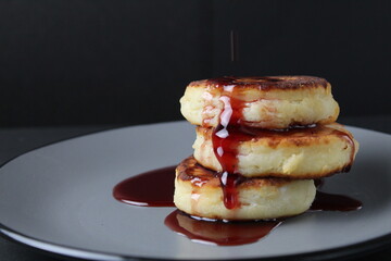 food breakfast cheesecakes curd pancakes three pieces lie stack of slides on top of each other and pour jam sauce cherry red on a gray plate on a black background