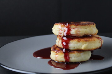 food breakfast cheesecakes curd pancakes three pieces lie stack of slides on top of each other and pour jam sauce cherry red on a gray plate on a black background