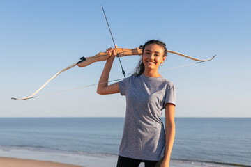 Smiling young woman holding bow and arrow while standing at beach