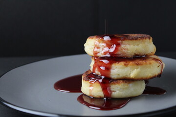 food breakfast cheesecakes curd pancakes three pieces lie stack of slides on top of each other and pour jam sauce cherry red on a gray plate on a black background