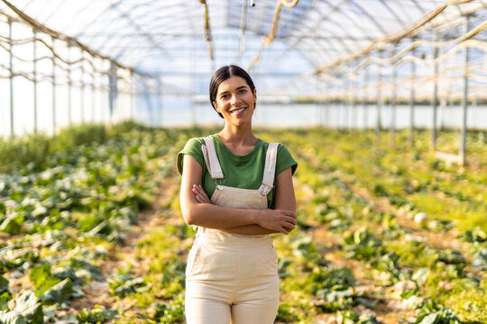 Smiling young female farm worker standing with arms crossed at greenhouse