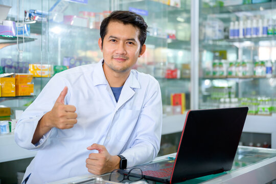 Young Male Pharmacist Smiling Happily In The Pharmacy Doing Inventory In A Well Stocked And Up-to-date Pharmacy. Pharmacist Using Computer At Pharmacy