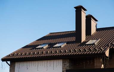 The roof is made of brown metal tiles against the blue sky. On the roof there is a house chimney and skylights.